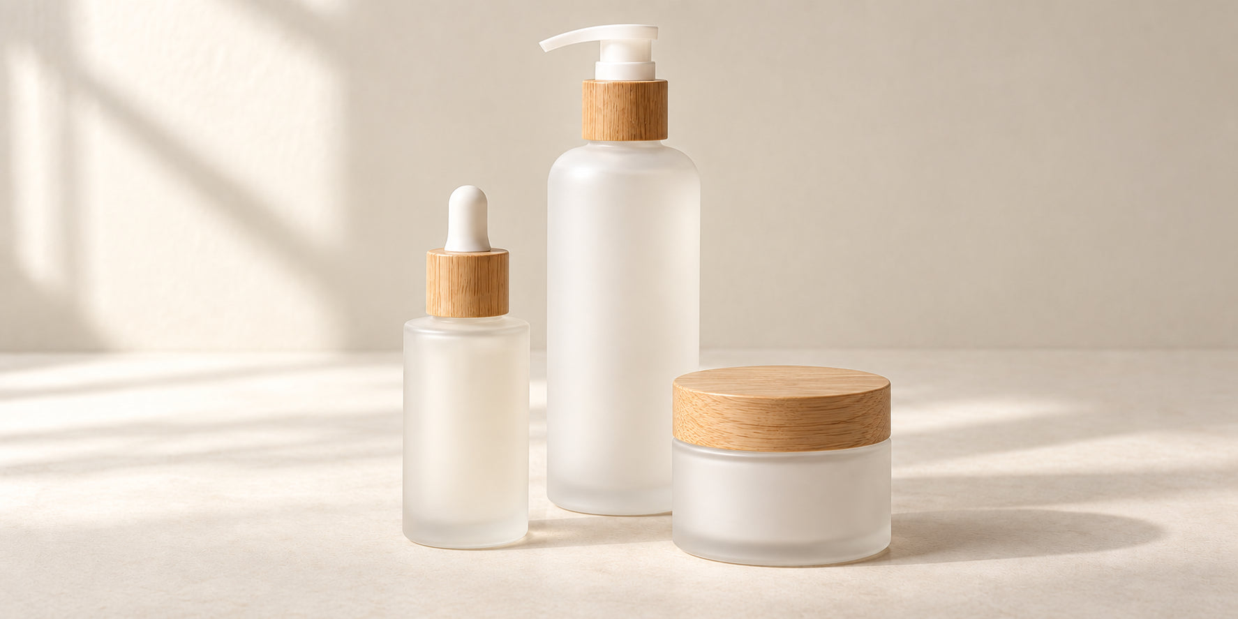 A frosted cleanser pump, serum bottle and closed cream jar with wood details in warm natural light